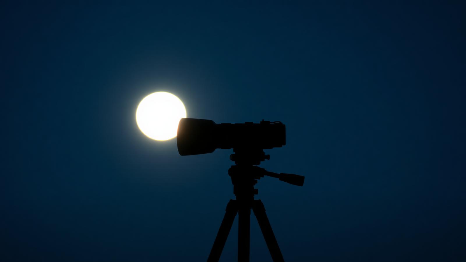 A large telephoto lens on a tripod silhouetted against a bright full moon