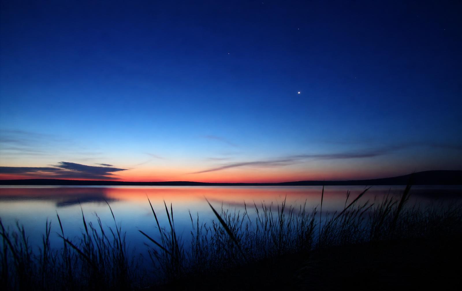 Deep blue nautical twilight over a still lake with faint stars appearing
